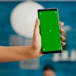 A hand holding a smartphone with a green screen against a blue background