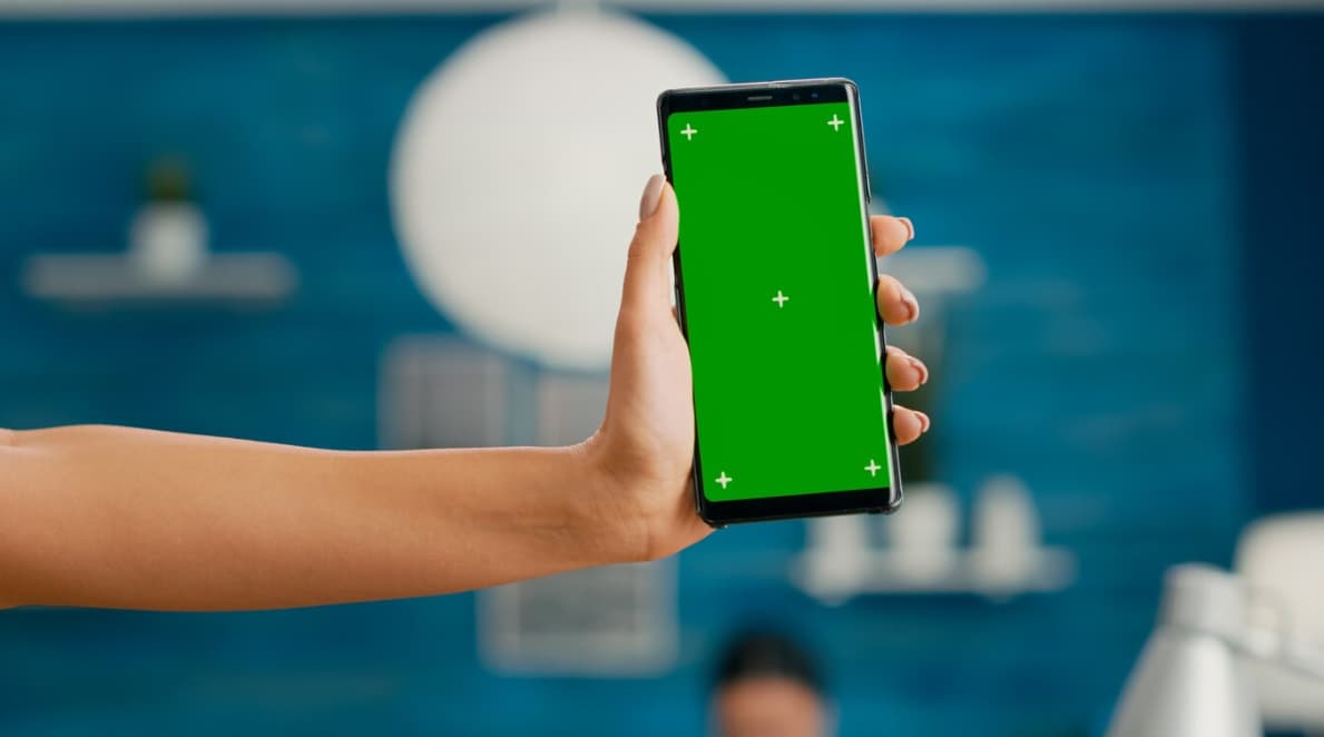 A hand holding a smartphone with a green screen against a blue background